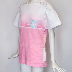 May include: A pink and white short-sleeved shirt with a graphic of a seagull flying over a sunset. The shirt has a white collar and a white panel on the back. The shirt has a white pocket on the right side.