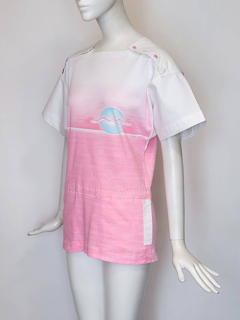 May include: A pink and white short-sleeved shirt with a graphic of a seagull flying over a sunset. The shirt has a white collar and a white panel on the back. The shirt has a white pocket on the right side.