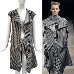 May include: A black and white houndstooth pattern vest with a unique asymmetrical design. The vest features a shawl collar and button details. The garment is displayed on a mannequin and a model, showcasing its distinctive style.