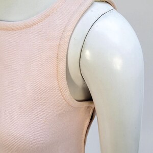 Puede incluir: Un top sin mangas de color rosa claro sobre un maniqu&iacute;. El top tiene una tela texturizada y un escote redondo. Las correas de los hombros son una extensi&oacute;n del escote. Se ve el brazo del maniqu&iacute;.