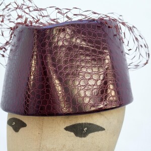 May include: A deep burgundy pillbox hat featuring a glossy, textured faux-alligator skin finish. The hat is adorned with a delicate, matching burgundy netting veil that extends over the top and sides of the structured, cylindrical crown.