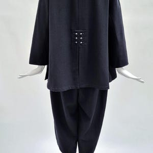 May include: A dark charcoal gray two-piece outfit consisting of a long-sleeved jacket and cropped trousers. The jacket features a notched collar and a back vent detailed with six small, round, silver-toned metallic buttons arranged in two rows.