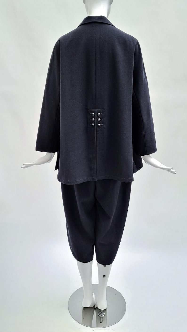 May include: A dark charcoal gray two-piece outfit consisting of a long-sleeved jacket and cropped trousers. The jacket features a notched collar and a back vent detailed with six small, round, silver-toned metallic buttons arranged in two rows.