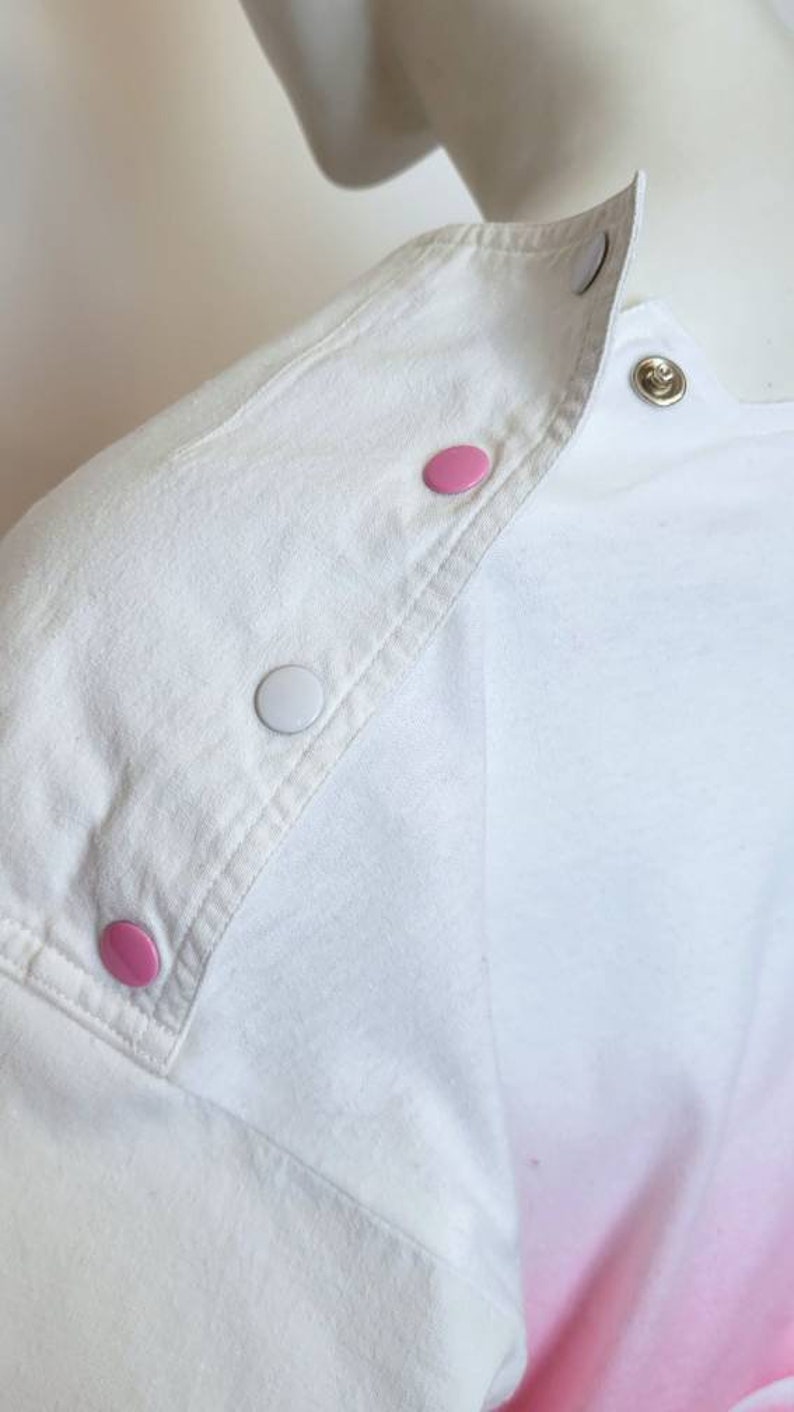 May include: White shirt with a pink and white ombre design. The shirt has a white collar and a row of pink and white buttons down the shoulder seam.