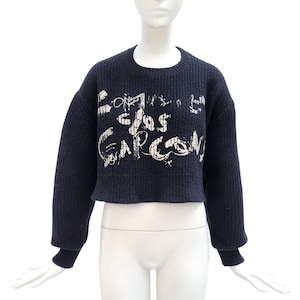 Puede incluir: Suéter corto azul marino con el texto "Comme des Garcons" en trazos de pincel blancos. El suéter tiene cuello redondo y mangas largas con puños acanalados. La prenda se exhibe en un maniquí.