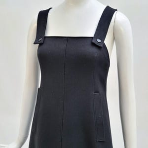 Pu&ograve; includere: Abito nero con ampie spalline e scollatura quadrata. L'abito presenta due tasche frontali e dettagli di bottoni sulle spalline. L'abito &egrave; esposto su un manichino.