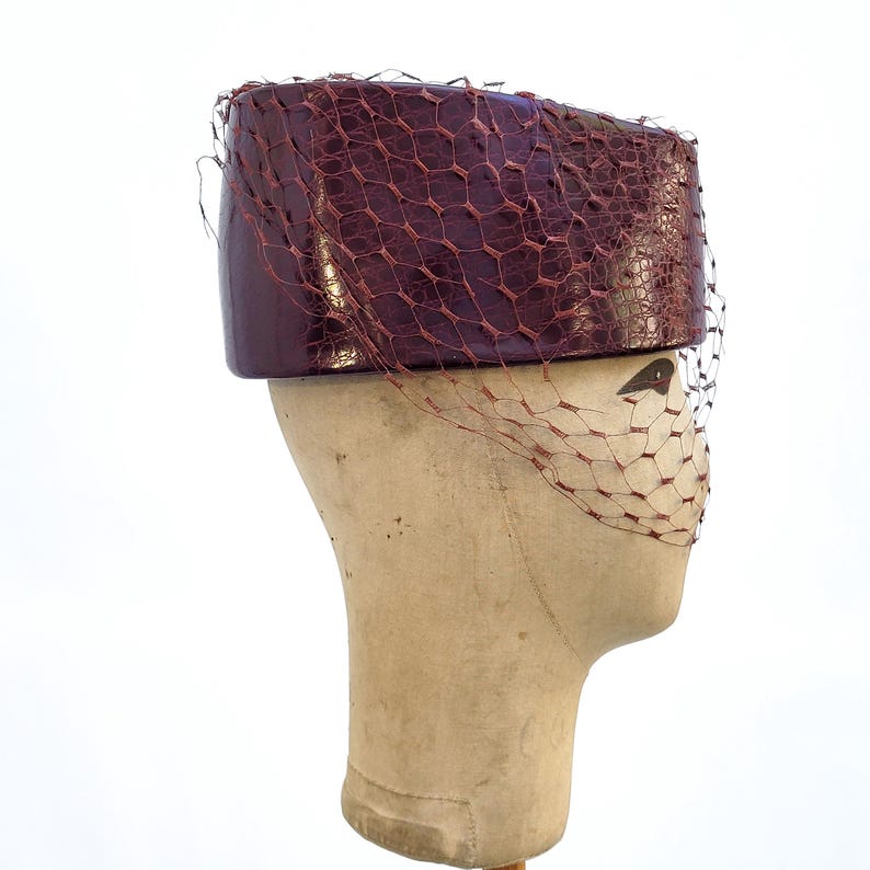 May include: A deep burgundy pillbox hat with a glossy, textured finish and a matching fine mesh netting veil that drapes over the front. The hat features a structured, cylindrical shape and is displayed on a neutral-toned mannequin head.