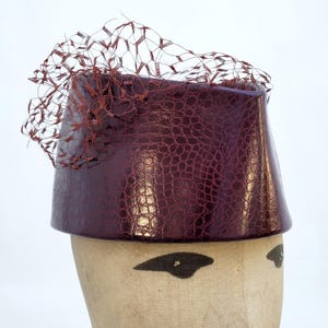 May include: A deep burgundy pillbox hat featuring a glossy, embossed crocodile skin texture. The hat is adorned with a matching dark red netting veil that extends upwards and outwards from the top edge, creating a structured and layered appearance.