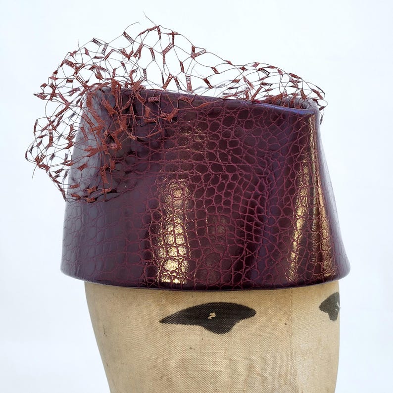 May include: A deep burgundy pillbox hat featuring a glossy, embossed crocodile skin texture. The hat is adorned with a matching dark red netting veil that extends upwards and outwards from the top edge, creating a structured and layered appearance.