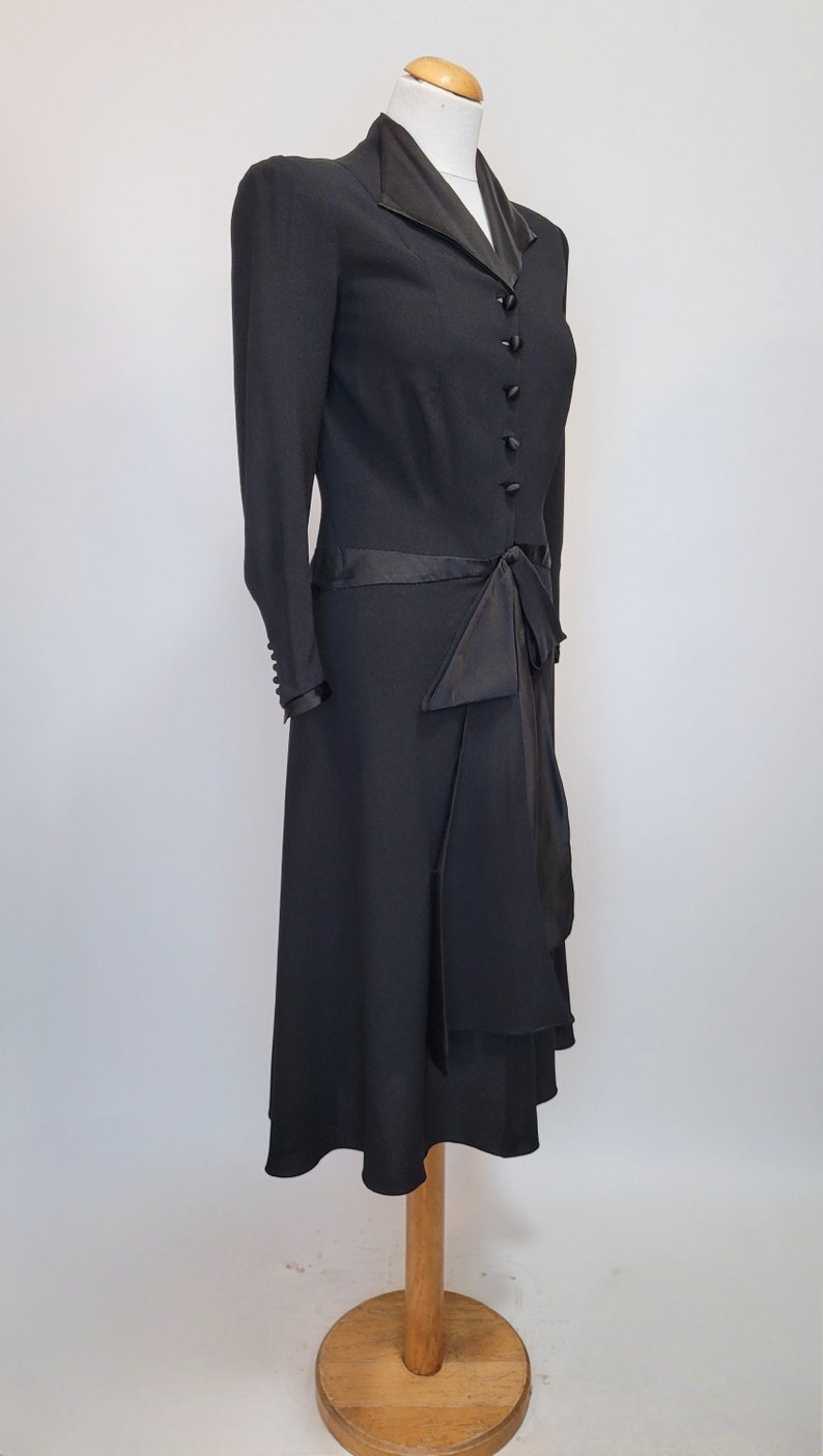 Puede incluir: Un vestido negro con cuello de sat&eacute;n y un lazo de sat&eacute;n en la cintura. El vestido tiene un corpi&ntilde;o ajustado y una falda fluida.