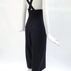 May include: A dark navy blue jumpsuit featuring a high-waisted design with wide-leg cropped trousers and thick shoulder straps that cross over the back, creating a clean, structured silhouette on a white mannequin display.