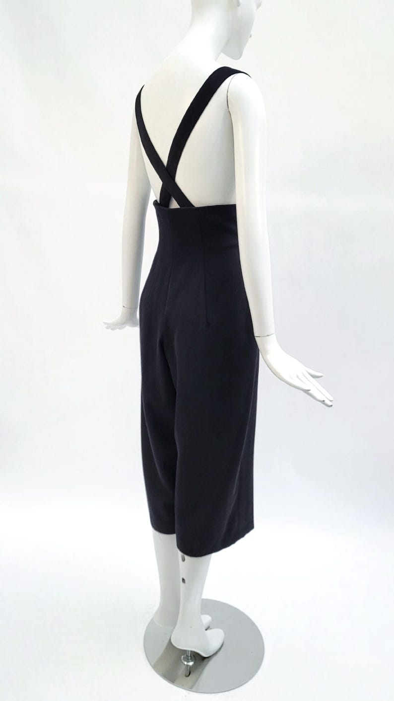 May include: A dark navy blue jumpsuit featuring a high-waisted design with wide-leg cropped trousers and thick shoulder straps that cross over the back, creating a clean, structured silhouette on a white mannequin display.