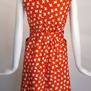 May include: A sleeveless orange dress with a white geometric pattern. The dress has a collared neckline, a belt, and pockets.