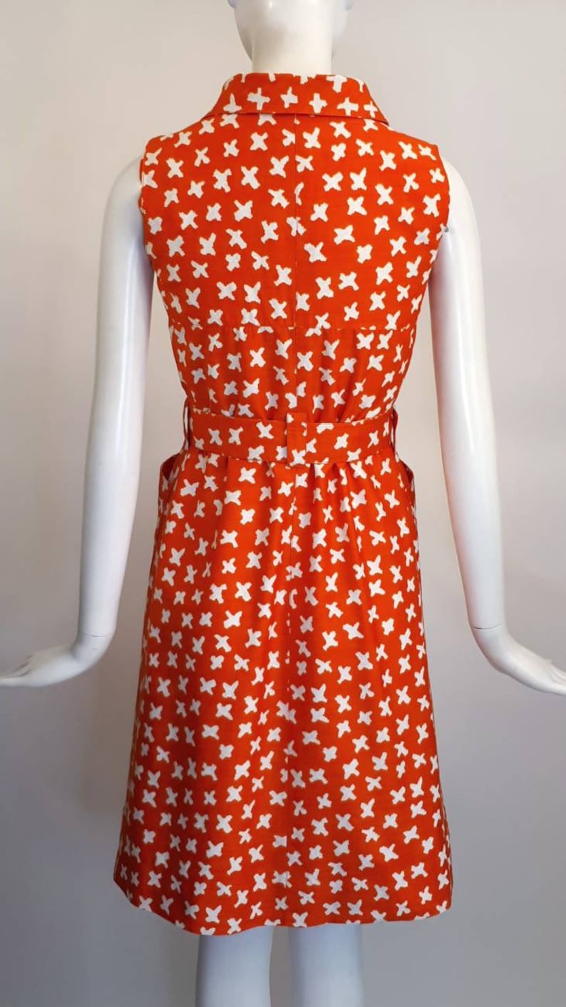 May include: A sleeveless orange dress with a white geometric pattern. The dress has a collared neckline, a belt, and pockets.