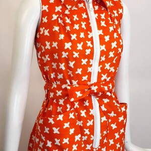 May include: A sleeveless orange dress with a white geometric pattern and a white zipper down the front. The dress has a white collar and a white belt.