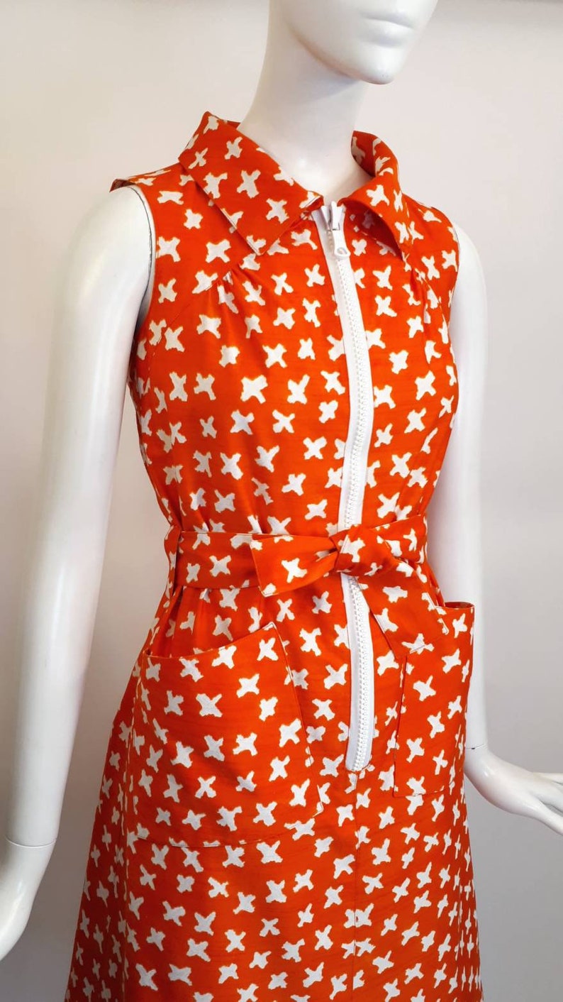 May include: A sleeveless orange dress with a white geometric pattern and a white zipper down the front. The dress has a white collar and a white belt.