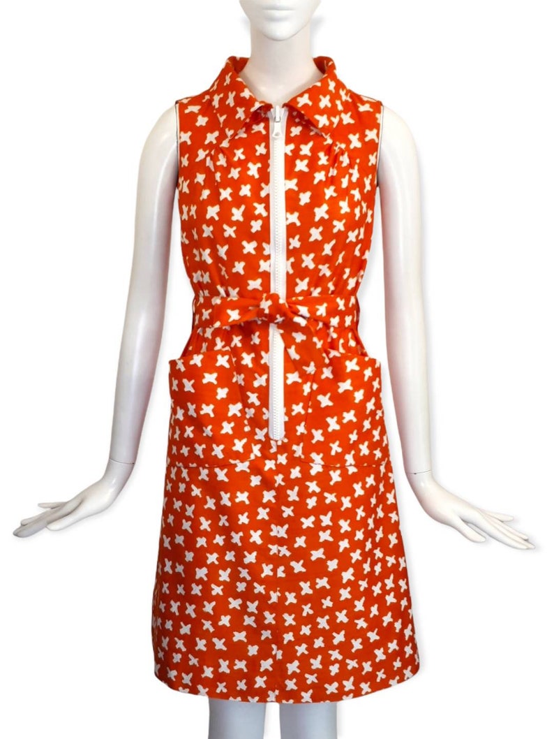 May include: A sleeveless orange dress with a white geometric pattern. The dress has a white collar, a zipper down the front, and a belt that ties at the waist.