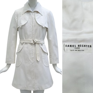 May include: Off-white trench coat with a matching belt and silver-tone buckle. The coat features a unique collar design, two front pockets, and metal ring details. The label inside reads "DANIEL HECHTER PARIS".