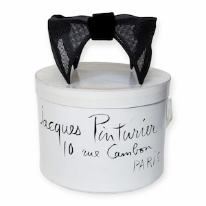 May include: A white hat box with black lettering that reads "Jacques Pinturier 10 rue Cambon PARIS". A black bow made of mesh fabric with a velvet centre sits on top of the box.