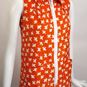 May include: A sleeveless orange dress with a white zipper down the front and a white geometric pattern. The dress has a collared neckline and two pockets.