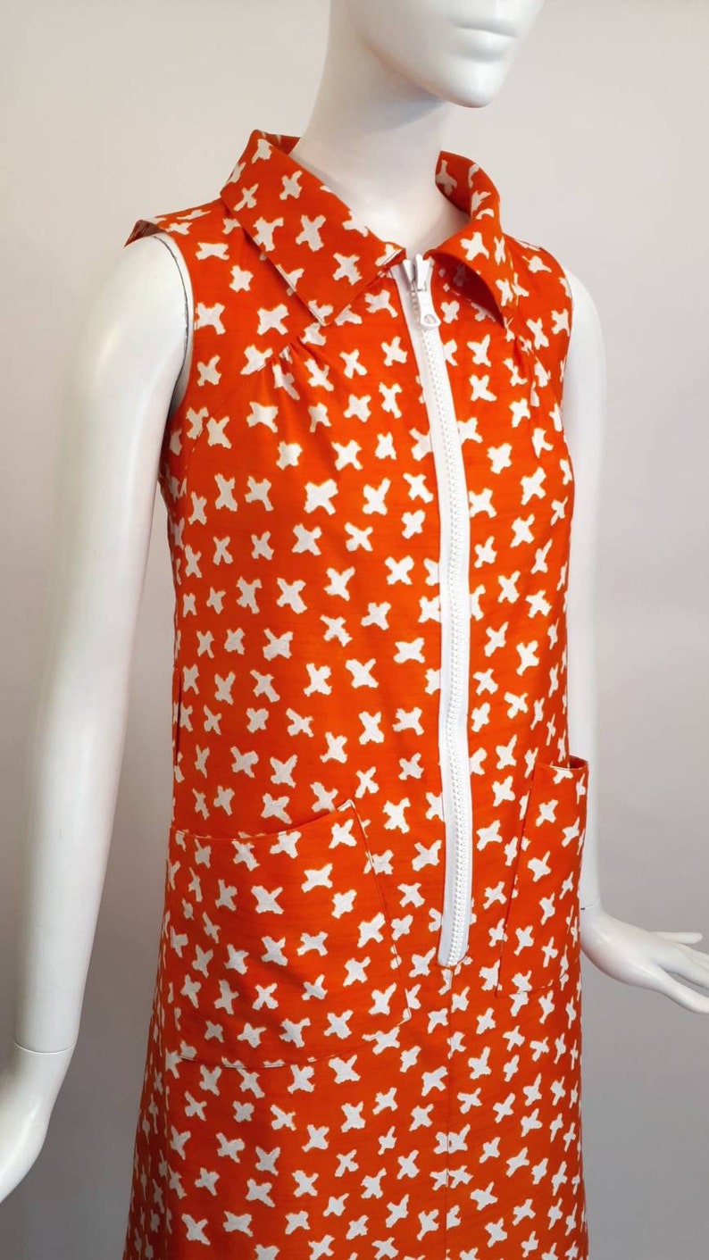 May include: A sleeveless orange dress with a white zipper down the front and a white geometric pattern. The dress has a collared neckline and two pockets.