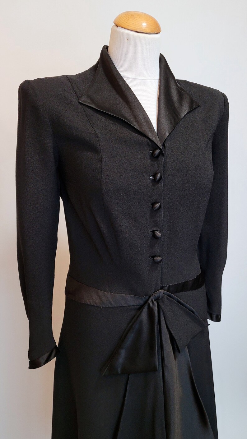 Puede incluir: Un vestido negro con cuello y lazo de sat&eacute;n. El vestido tiene mangas largas y un corpi&ntilde;o ajustado. El vestido se abrocha con botones en la parte delantera.