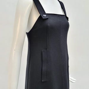 Pu&ograve; includere: Abito nero senza maniche con ampie spalline e una tasca rettangolare. Le spalline sono fissate con un bottone. L'abito &egrave; esposto su un manichino.
