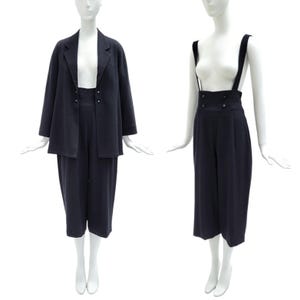 May include: A dark navy blue two-piece set consisting of a long-sleeved blazer and high-waisted cropped trousers. The trousers feature wide shoulder straps and two rows of small buttons at the waist. The blazer has a notched lapel collar.