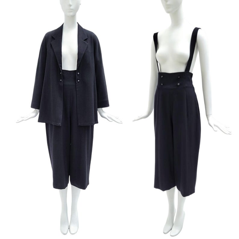 May include: A dark navy blue two-piece set consisting of a long-sleeved blazer and high-waisted cropped trousers. The trousers feature wide shoulder straps and two rows of small buttons at the waist. The blazer has a notched lapel collar.