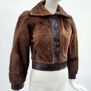 May include: A brown faux fur jacket with a dark brown leather collar, trim, and button closure. The jacket has long sleeves and a cropped length, displayed on a mannequin. The jacket's texture is soft and fluffy.