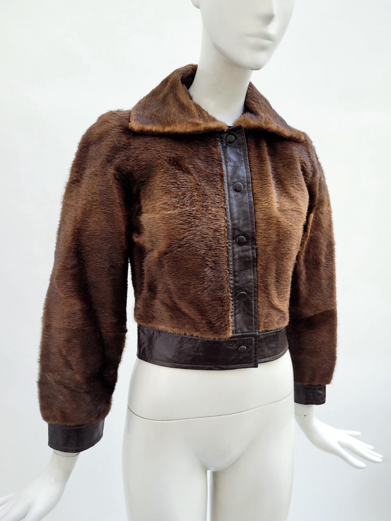 May include: A brown faux fur jacket with a dark brown leather collar, trim, and button closure. The jacket has long sleeves and a cropped length, displayed on a mannequin. The jacket's texture is soft and fluffy.