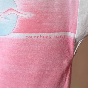 May include: A pink and white t-shirt with a graphic of a woman swimming in a circle. The text "courr&egrave;ges paris" is printed on the shirt.