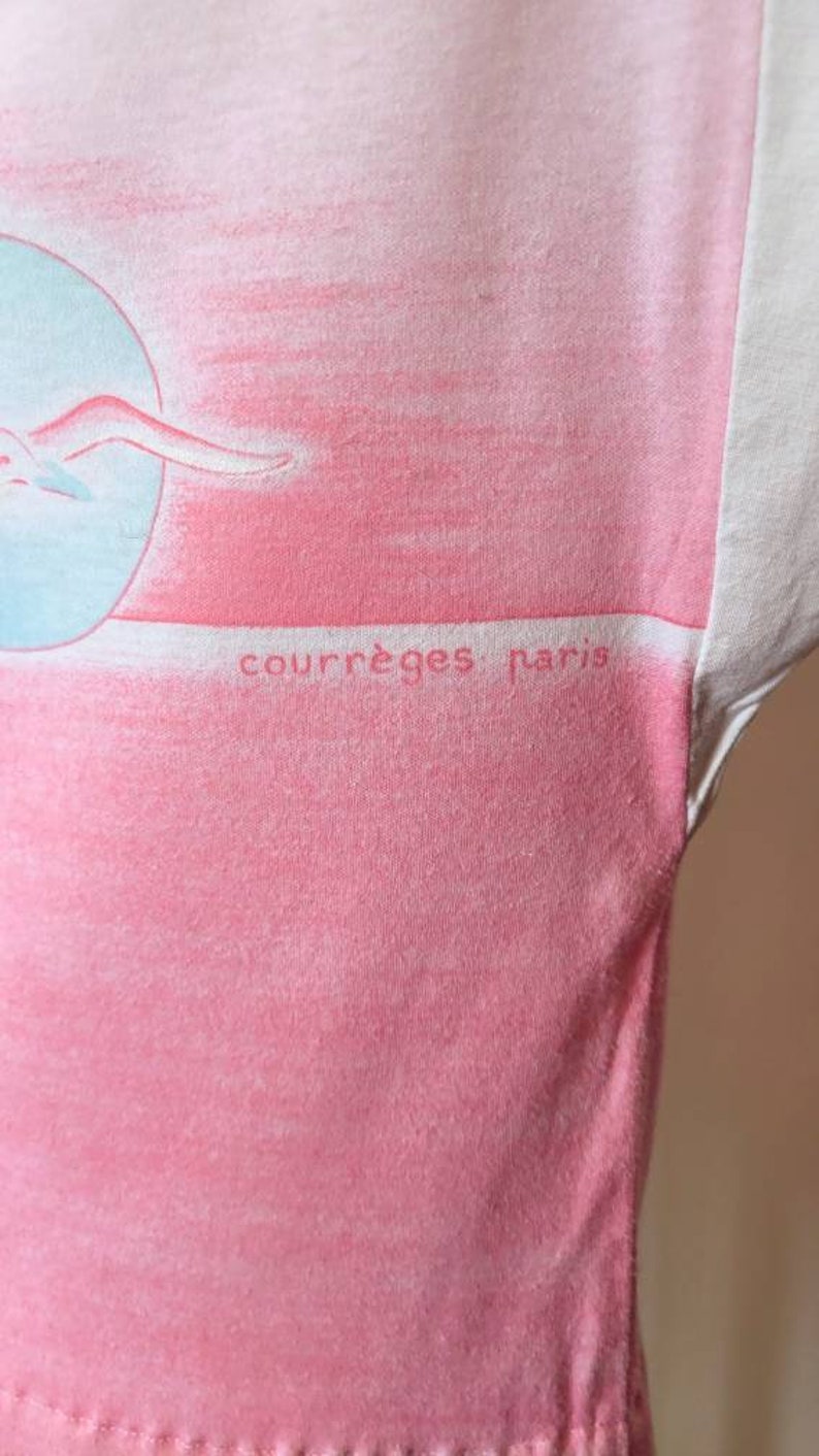 May include: A pink and white t-shirt with a graphic of a woman swimming in a circle. The text "courr&egrave;ges paris" is printed on the shirt.