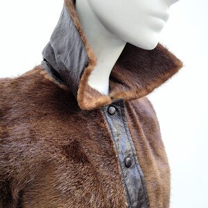 May include: A brown fur coat with a dark brown leather collar and trim. The coat features a button-up front and is displayed on a mannequin. The fur has a soft, luxurious appearance, with a rich, warm tone.