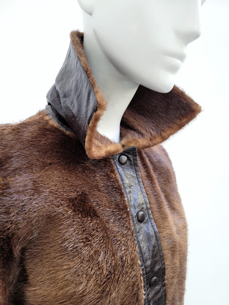 May include: A brown fur coat with a dark brown leather collar and trim. The coat features a button-up front and is displayed on a mannequin. The fur has a soft, luxurious appearance, with a rich, warm tone.