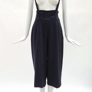 May include: A pair of dark navy blue high-waisted trousers with attached suspender straps. The trousers feature a wide waistband with four small buttons arranged in two rows at the front and a cropped, wide-leg silhouette that falls below the knee.