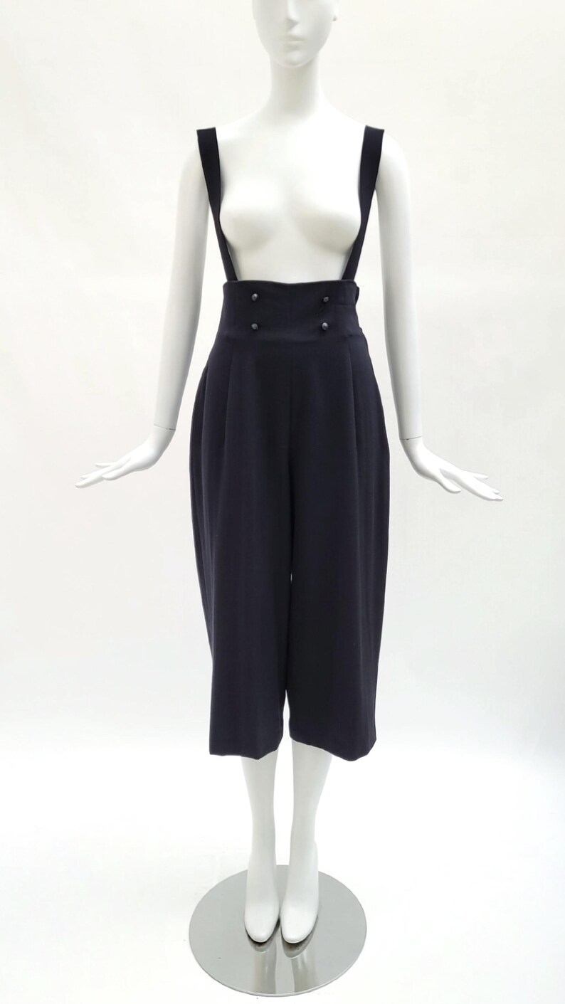 May include: A pair of dark navy blue high-waisted trousers with attached suspender straps. The trousers feature a wide waistband with four small buttons arranged in two rows at the front and a cropped, wide-leg silhouette that falls below the knee.