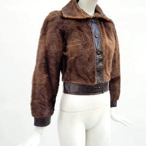 May include: A brown fur jacket with a dark brown leather collar, button placket, and cuffs. The jacket is displayed on a mannequin. The jacket's waist is cinched with a dark brown leather band.