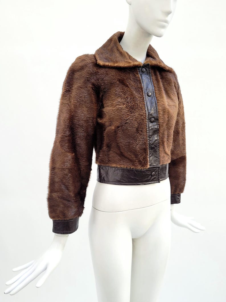 May include: A brown fur jacket with a dark brown leather collar, button placket, and cuffs. The jacket is displayed on a mannequin. The jacket's waist is cinched with a dark brown leather band.