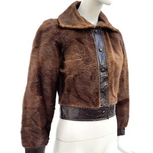 May include: A brown faux fur jacket with dark brown leather trim and button closure. The jacket features a collar and long sleeves with leather cuffs. The jacket is displayed on a mannequin.