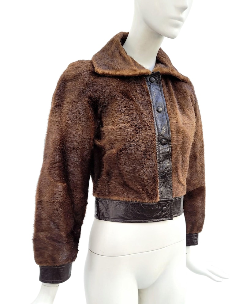 May include: A brown faux fur jacket with dark brown leather trim and button closure. The jacket features a collar and long sleeves with leather cuffs. The jacket is displayed on a mannequin.