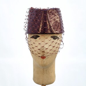 May include: A cylindrical pillbox hat in a deep burgundy, textured faux-leather finish. The hat features a matching burgundy netting veil that drapes over the front, creating a structured and layered appearance with a distinct hexagonal mesh pattern.