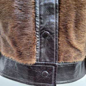 May include: Close-up of a brown jacket with a fur texture and dark brown leather accents. The jacket features a button-up front and a structured design. The fur has a natural, textured appearance, and the leather has a glossy finish.