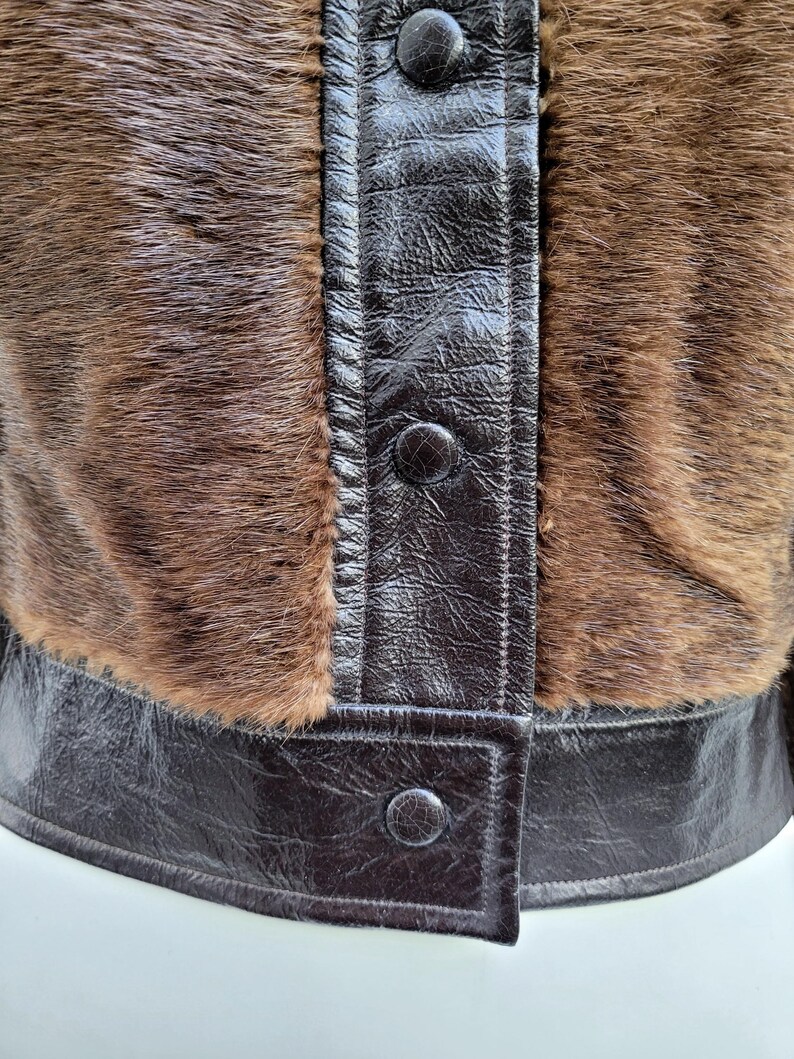 May include: Close-up of a brown jacket with a fur texture and dark brown leather accents. The jacket features a button-up front and a structured design. The fur has a natural, textured appearance, and the leather has a glossy finish.