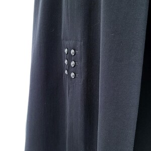 May include: A dark charcoal gray garment featuring a vertical panel with six small, round, matching buttons arranged in two parallel columns of three. The fabric has a smooth, matte texture and a clean, straight hemline along the bottom edge.