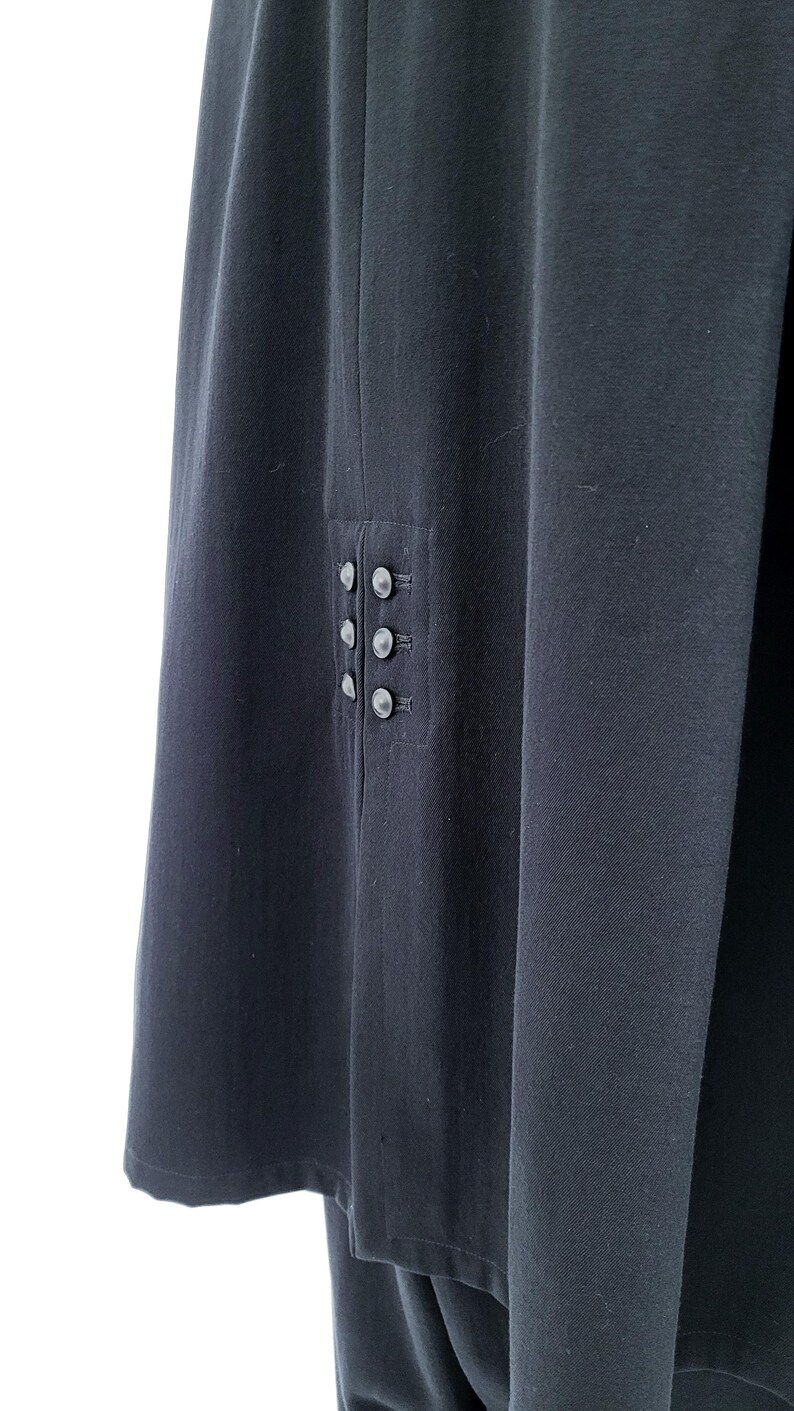 May include: A dark charcoal gray garment featuring a vertical panel with six small, round, matching buttons arranged in two parallel columns of three. The fabric has a smooth, matte texture and a clean, straight hemline along the bottom edge.
