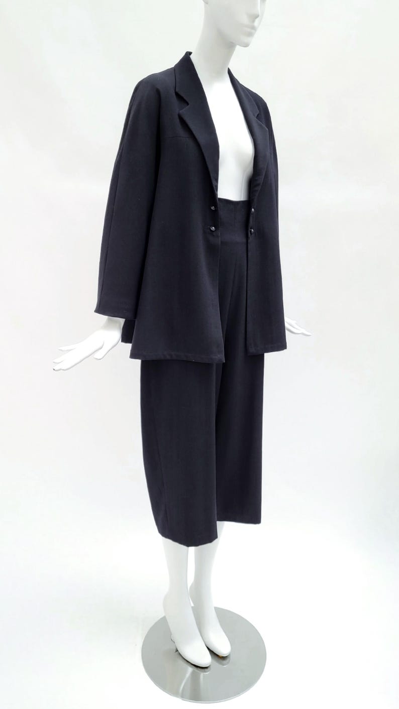 May include: A dark navy blue two-piece set consisting of a long-sleeved blazer with a notched lapel and two front buttons, paired with matching wide-leg cropped trousers. The ensemble is displayed on a white mannequin against a plain background.