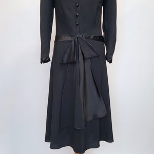 Puede incluir: Un vestido negro con cuello de sat&eacute;n y un lazo de sat&eacute;n en la cintura. El vestido tiene mangas largas y una falda hasta la rodilla.