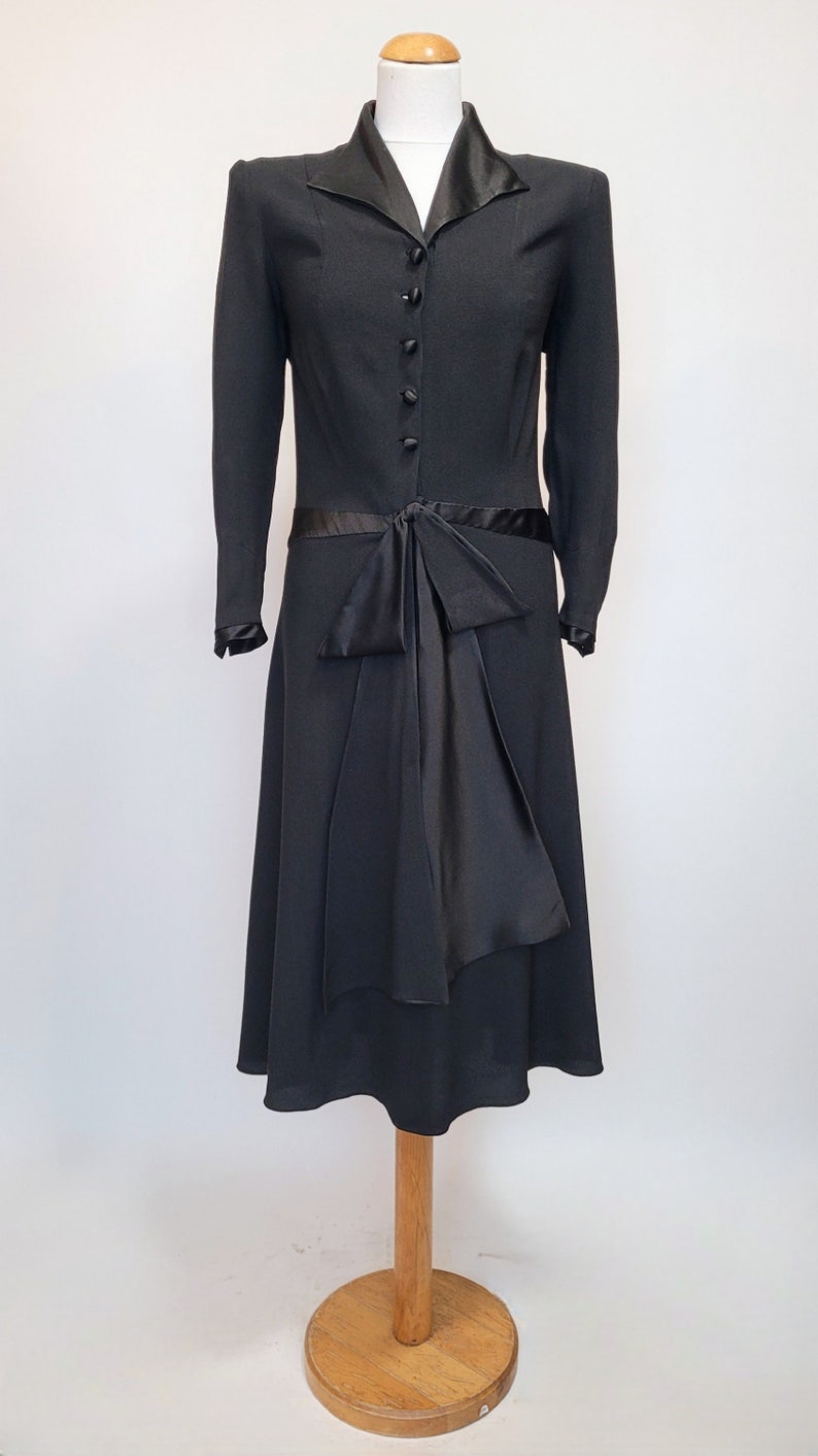 Puede incluir: Un vestido negro con cuello de sat&eacute;n y un lazo de sat&eacute;n en la cintura. El vestido tiene mangas largas y una falda hasta la rodilla.