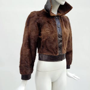 May include: A brown fur jacket with a high collar and leather detailing. The jacket has a dark brown leather collar, button-up front, and cuffs. The waistline and cuffs are trimmed with dark brown leather. The jacket is displayed on a mannequin.